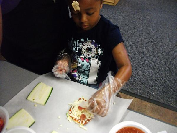 A child wearing plastic gloves places cheese and other pizza ingredients on a slice of eggplant.